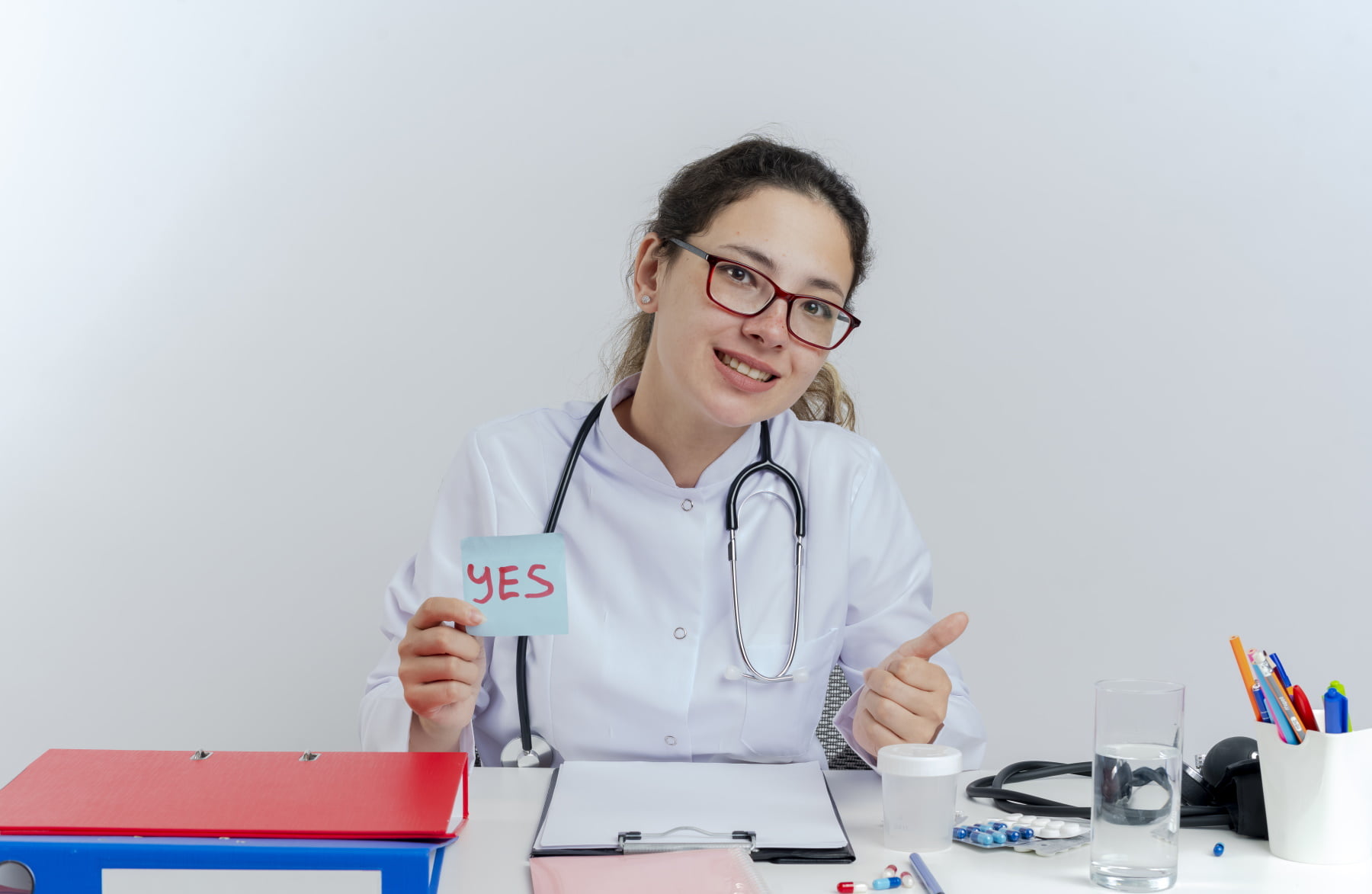 smiling young female doctor wearing medical robe and stethoscope and glasses sitting at desk with medical tools looking at camera holding yes note showing thumb up isolated on white background