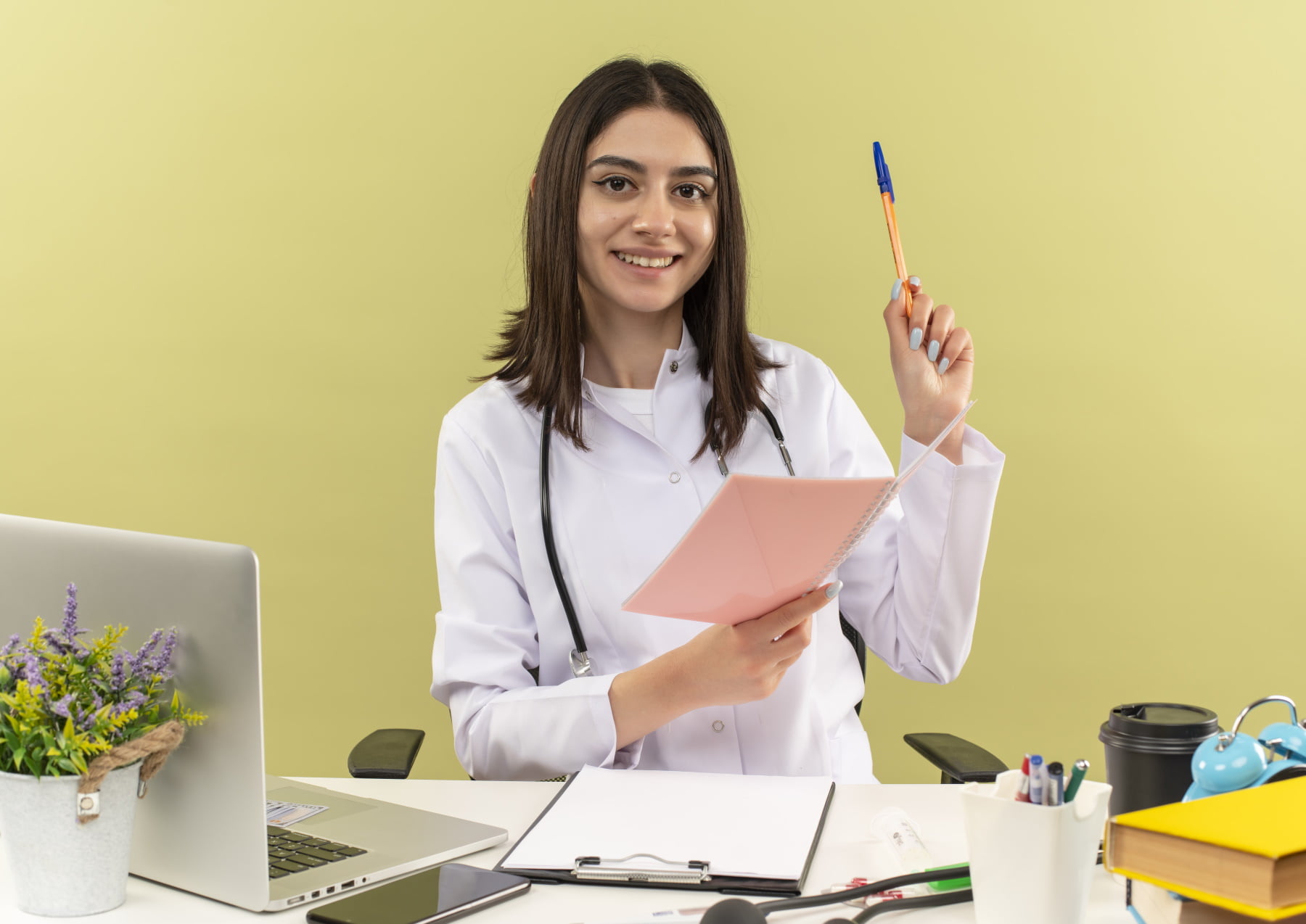 young woman doctor in white coat with stethoscope around her neck holding notebook and pen looking at camera wirh smile on face sitting at the table with laptop over light background